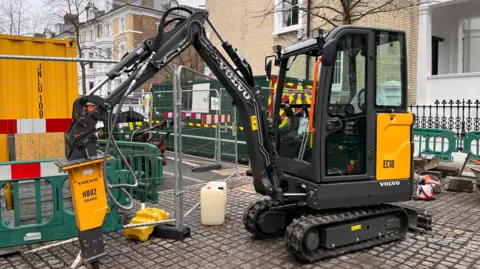 Transport for London A yellow and black Volvo electric excavator on a road surrounded by barriers and metal-corrugated green and yellow portable cabins.