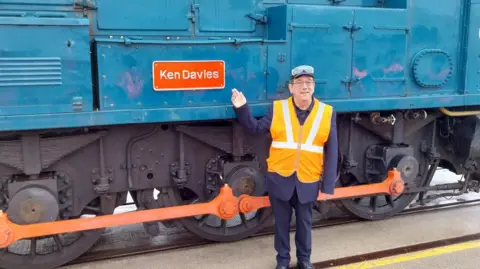 Alstom Ken Davies standing beside the train which bares his name, pointing to the plaque. He is wearing a high vis orange jacket, a navy suit, and goggles upon his head.