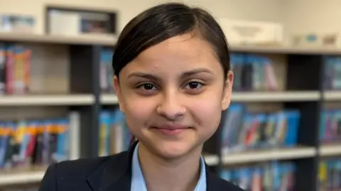 A close-up headshot of a young girl smiling into the camera. Her hair is tied back with side parting. She has brown eyes and is wearing a blue shirt and navy blazer.