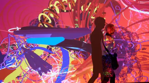 Andy Willsher A person walking past a virtual reality video on a wall in an art gallery. The colours a bright, bold and colourful. 