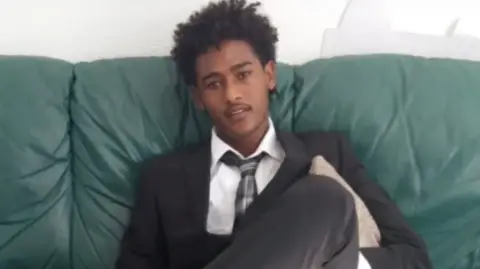 Family A young man sits on a green leather sofa wearing a shirt, tie and suit. He looks directly at the camera.