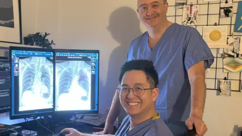 Harrogate and District NHS Foundation Trust Consultant radiologists Dr Daniel Fascia and Dr Andrew Koo, who wear blue scrubs, sit and stand next to a computer showing a chest x-ray.