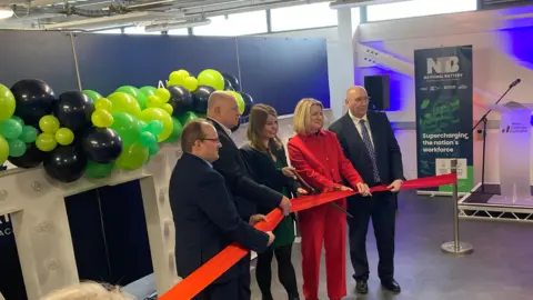 BBC Five people holding a red ribbon as part of an opening ceremony. Left to right: a partially bald man in a dark suit, wearing spectacles; a bald man in a dark suit; a smiling woman with shoulder-length brown hair, wearing a dark green dress and black jacket, holding a pair of shears and about to cut the ribbon; a woman with blonde shoulder-length hair wearing a red suit, and a balding man wearing spectacles, a dark suit and a blue tie. Behind them is a strand of green and black balloons