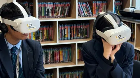 BBC Two students, one boy and one girl sitting with virtual reality headset on. A bookshelf is behind them. They are wearing navy blazers and blue, tie with green stripes.