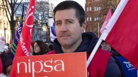 BBC Ryan Wilson with short, dark hair looks into the camera at a Nipsa picket line. He is holding a red Nipsa sign and a red flag. He is surrounded by people who are also holding red and blue Nipsa flags.