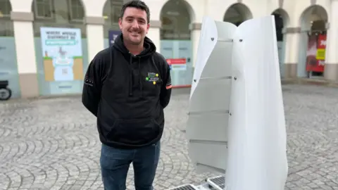 BBC A man stands next to a white wind turbine, which is made of two curved, vertical sails on a flat base. The turbine is slightly shorter than the man.