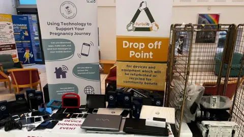 South Tees Hospitals NHS Foundation Trust A table containing a number of electronic devices - mostly laptops and mobile phones. It is backed by a sign saying "Drop off Point for devices and equipment which will be refurbished or recycled for community benefit" and another stating "Using Technology to Support your Pregnancy Journey".
