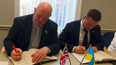 BBC A photograph of Councillor Colin Davie and Oleksandr Prokudin bent over a desk, both signing documents of the agreement