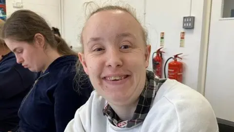 BBC Jessica has a white jumper and checkered shirt. She has light coloured hair and there is a woman in a blue jumper behind her. They are both in a white walled room