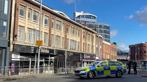 BBC A police car is parked across a road. Two police officers in black clothes stand behind the vehicle looking away from the camera, police tape is behind them.