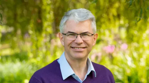 University of Surrey A man in a purple jumper with a blue shirt underneath, he is wearing glasses and has grey hair. The background is blurred but he is outside next to some green plants.