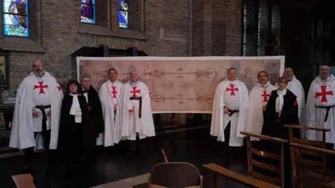 Brother John Michael A group of people, some dressed in white robes with red crosses, stand inside a church in front of a large replica of the Shroud of Turin. The church features stained-glass windows and wooden chairs.