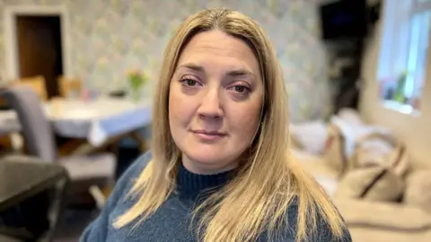 NIKKI FOX/BBC Sarra Wilson looks at the camera. She is sitting in her open plan kitchen which has been adapted for wheelchair use. She has long blonde hair and is wearing a green wool jumper.