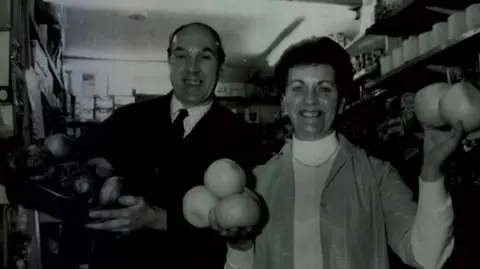 Family A black and white photo of a man, on the left, and a woman, on the right, holding up fruit and veg and grinning. They are in a shop, surrounded by shelving. He is wearing an overcoat, checked shirt and tie, and she has an over-shirt over a polo-neck.