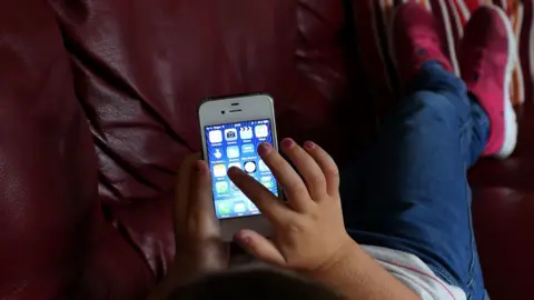 PA An over the shoulder shot of a child as they scroll through the home screen of an iPhone 4 while lying on a sofa.They are wearing a white top with thin red stripes, blue jeans, and pink trainers with white soles.
