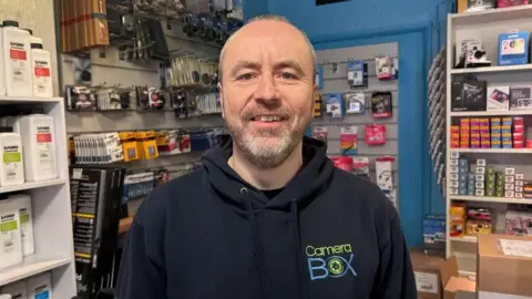 Kate Bradbrook/BBC Adrian Clarke with very short dark hair and beard, wearing a Camera Box hoodie and standing in his shop. Shelves are visible with boxes of film, smart cards, cameras and accessories behind him.
