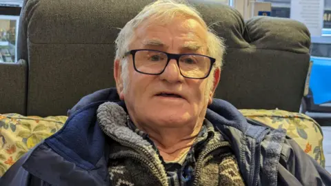BBC A man wearing glasses, a navy blue coat and a pullover sitting on a yellow and blue patterned sofa.