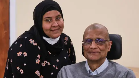 Peterborough City Council A younger woman (on the left) is standing and smiling next to an older man (on the right), who has a head rest behind him. The woman is smiling and wearing a black top with pink flowers and a black hijab. The man is smiling and wearing glasses, a light coloured shirt and a grey jumper. Behind them is a plain wall.